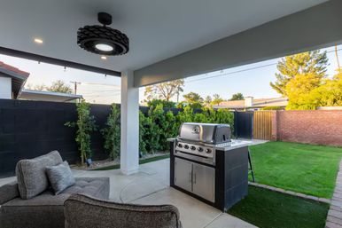 Cozy modern suburban covered backyard patio with stainless steel gas grill, gray cushioned outdoor sofa, overhead light fixture, neatly trimmed hedges, green lawn, black privacy wall and wooden gate