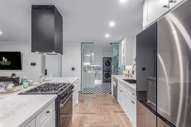 Modern white kitchen with marble countertops and island gas range under a black vent hood, stainless steel refrigerator, herringbone wood-look tile floor, and glass-doored laundry area with black-and-white checkered floor and stacked washer-dryer.