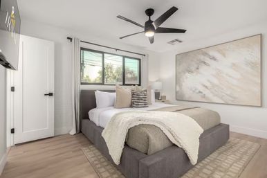 Bright modern master bedroom with gray upholstered platform bed, neutral bedding and knit throw, abstract wall art, black ceiling fan and wood floors