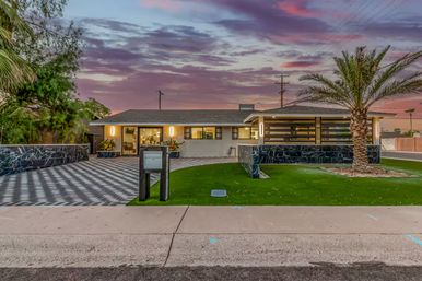 Mid-century modern single-story suburban home with patterned driveway, marble accent walls, palm tree and vibrant sunset sky.