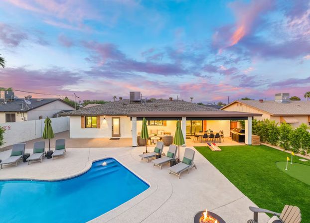 Suburban backyard at sunset: blue swimming pool with lounge chairs and green umbrellas, covered patio with outdoor dining and grill, cornhole boards, a small putting green and a glowing fire pit.