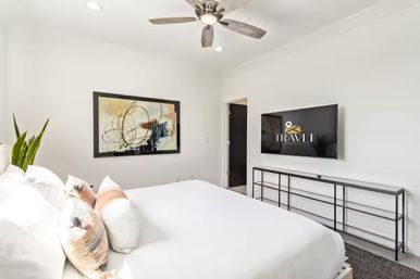 Bright modern bedroom interior with white walls, king bed and decorative pillows, abstract wall art, ceiling fan, wall-mounted TV and slim black console — stylish vacation rental vibe.