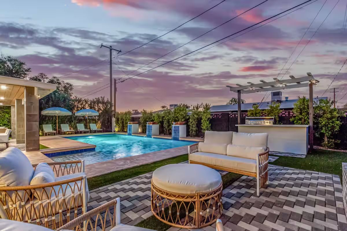 Twilight residential backyard with illuminated swimming pool, cushioned rattan patio seating and ottoman, pergola bar, poolside loungers with umbrellas and colorful sunset sky.