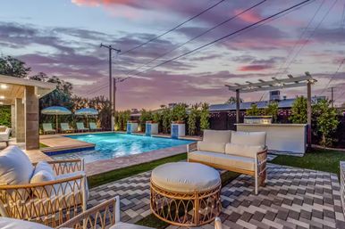 Twilight residential backyard with illuminated swimming pool, cushioned rattan patio seating and ottoman, pergola bar, poolside loungers with umbrellas and colorful sunset sky.