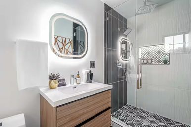 Bright, stylish modern bathroom with wood vanity and white sink, LED-lit rounded mirror, glass walk-in shower with black accent tiles, rainfall head and geometric floor tiles