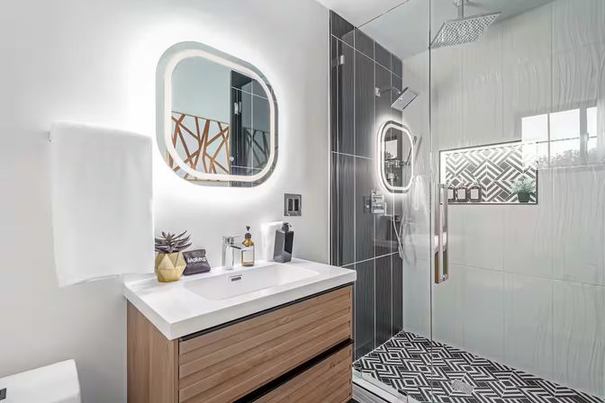 Bright, stylish modern bathroom with wood vanity and white sink, LED-lit rounded mirror, glass walk-in shower with black accent tiles, rainfall head and geometric floor tiles