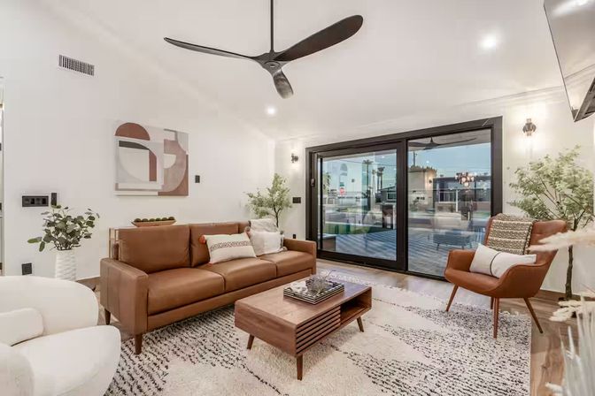 Bright modern living room with a brown leather sofa, mid-century wooden coffee table and matching armchair on a textured rug, sleek black ceiling fan and large sliding glass doors opening to a patio with potted plants.
