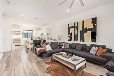 Sunlit modern open-concept living room and kitchen with a large charcoal sectional, white coffee table with chess set, abstract black-and-gold wall art, and light wood-look tile flooring.