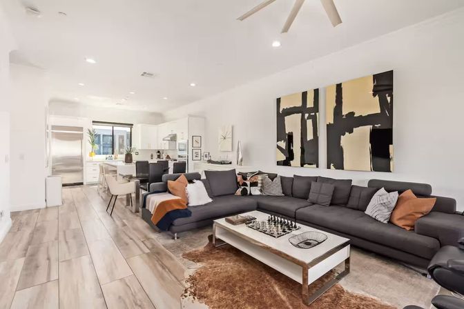 Sunlit modern open-concept living room and kitchen with a large charcoal sectional, white coffee table with chess set, abstract black-and-gold wall art, and light wood-look tile flooring.