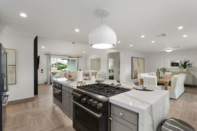 Bright open-concept kitchen in a suburban home featuring a marble waterfall island with gas range, gray cabinetry, large white pendant, hardwood-look floors, and adjacent dining and living areas with sliding glass door.