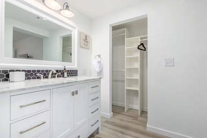 Bright modern white bathroom with marble countertop vanity, hexagon black tile backsplash, large framed mirror and wall lights, and an open walk-in closet with built-in white shelving, wood-look floors and a single black hanger.