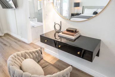 Contemporary bedroom nook with a floating glossy black vanity with gold knobs beneath a round mirror, stacked books and a decorative box, plush tufted chair and light hardwood floor near a bathroom doorway.
