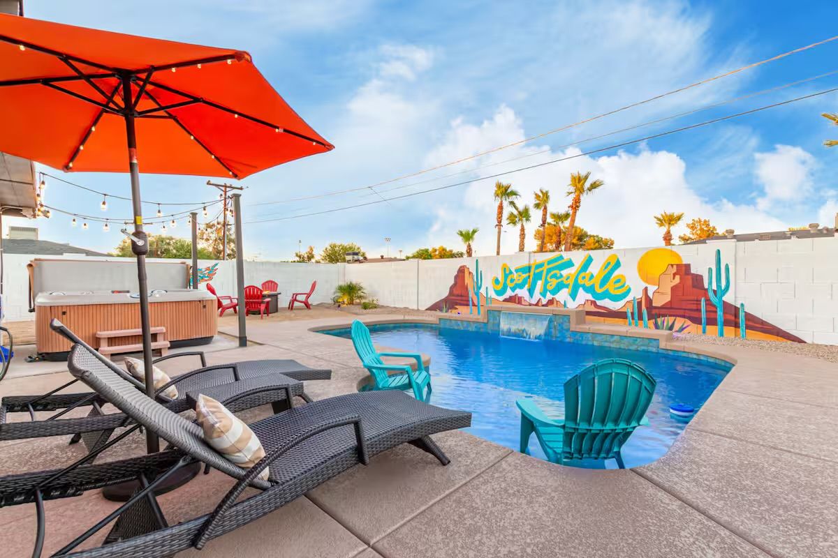 Bright Scottsdale backyard pool scene with turquoise Adirondack chairs partially in the water, wicker lounge loungers under an orange umbrella, a hot tub, red patio chairs, and a colorful desert mural on a white block wall under a blue sky.