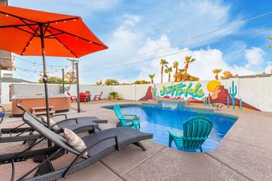 Bright Scottsdale backyard pool scene with turquoise Adirondack chairs partially in the water, wicker lounge loungers under an orange umbrella, a hot tub, red patio chairs, and a colorful desert mural on a white block wall under a blue sky.