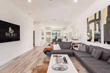 Stylish open-concept living room and kitchen with a large gray sectional sofa, colorful throw pillows, a coffee table with a chessboard, light wood floors, abstract wall art, and a wall-mounted TV in a bright minimalist space.
