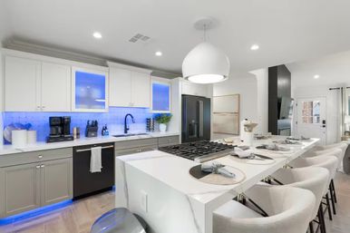 Bright modern open-concept kitchen with white quartz waterfall island and gas cooktop, plush bar stools, stainless refrigerator, and blue LED under-cabinet lighting