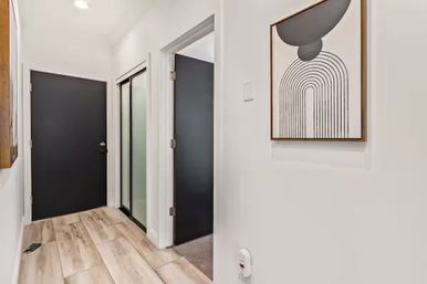 Modern apartment hallway with light wood-look tile floor, matte black entry and interior doors, frosted glass closet doors, and framed abstract arch wall art.