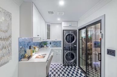 Bright modern laundry room with stacked washer-dryer, white cabinetry, blue scallop tile backsplash, built-in desk and geometric black-and-white floor tiles
