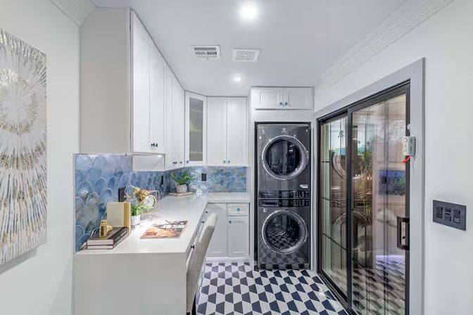 Bright modern laundry room with stacked washer-dryer, white cabinetry, blue scallop tile backsplash, built-in desk and geometric black-and-white floor tiles