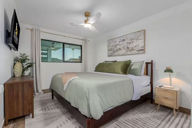 Serene modern bedroom with sage-green bedding on a wooden bed frame, wall-mounted TV, wooden dresser, bedside table with lamp, abstract wall art, ceiling fan and window with curtains.