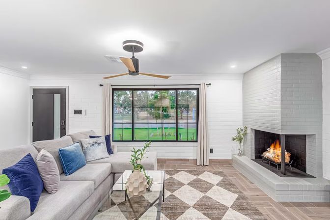 Bright modern suburban living room with light-gray sectional and blue pillows, glass coffee table, patterned rug, large black-framed window overlooking a green yard, white painted brick fireplace with a roaring fire, and a wood-blade ceiling fan.