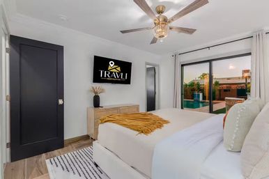 Bright modern bedroom with king bed, mustard throw and patterned pillows, wall-mounted flat-screen TV, wooden dresser with vase, ceiling fan, and sliding glass doors opening to a backyard pool patio.
