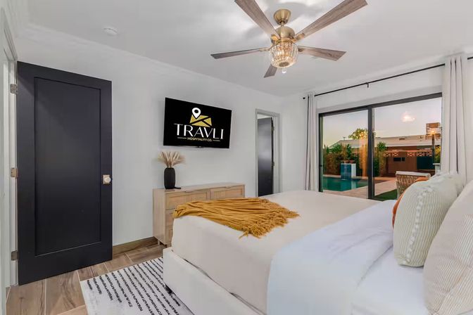 Bright modern bedroom with king bed, mustard throw and patterned pillows, wall-mounted flat-screen TV, wooden dresser with vase, ceiling fan, and sliding glass doors opening to a backyard pool patio.