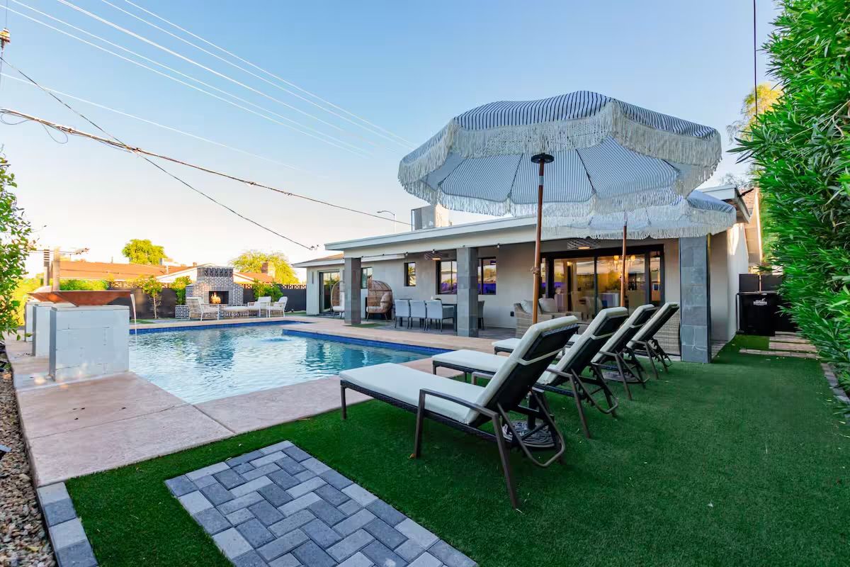 Sun-ready modern backyard with a rectangular swimming pool, white lounge chairs on artificial turf under striped fringe umbrellas, a covered patio dining area, and an outdoor fireplace.