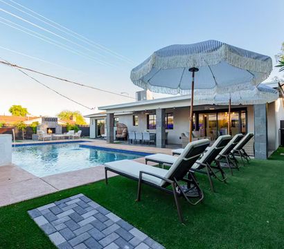 Sun-ready modern backyard with a rectangular swimming pool, white lounge chairs on artificial turf under striped fringe umbrellas, a covered patio dining area, and an outdoor fireplace.