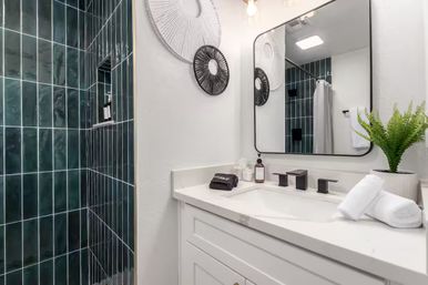 Modern bathroom with deep green vertical subway tiles in shower, white vanity with quartz countertop, matte-black faucet and framed mirror, potted fern and rolled towels.