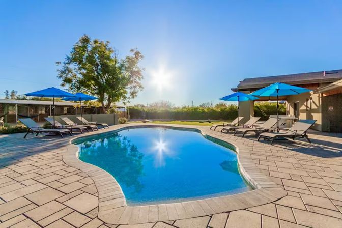 Resort-style outdoor swimming pool with sparkling blue water, bright sun reflection, stone patio, lounge chairs and blue umbrellas in a landscaped backyard