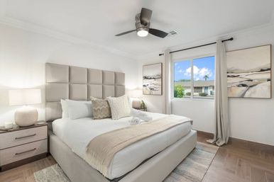 Bright modern master bedroom with tufted beige headboard, plush white bedding and textured throw pillows, matching nightstands and lamps, ceiling fan, and a window framing a sunny suburban street and blue sky.