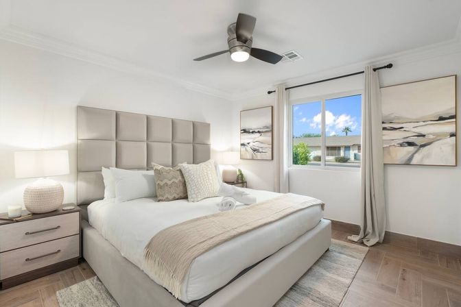 Bright modern master bedroom with tufted beige headboard, plush white bedding and textured throw pillows, matching nightstands and lamps, ceiling fan, and a window framing a sunny suburban street and blue sky.