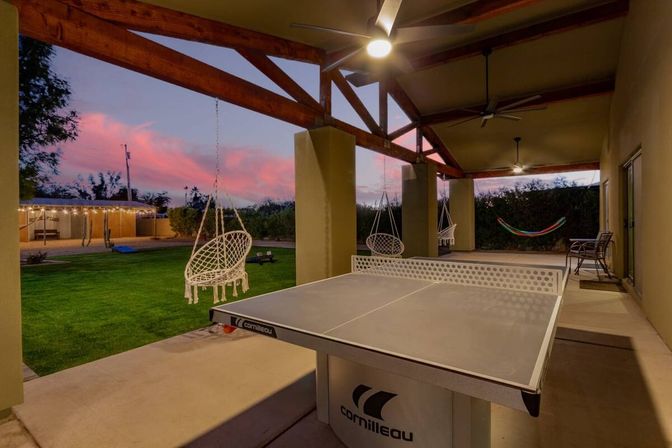 Covered backyard patio at sunset with a ping-pong table, hanging macramé swing chairs, hammock and green lawn — fun outdoor entertainment area
