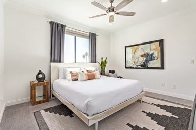 Sunny modern bedroom with white platform bed and crisp linens, wooden bedside table, abstract wall art, ceiling fan, gray curtains, potted plant, and palm trees visible outside the window.
