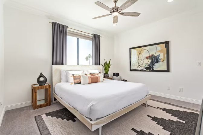 Sunny modern bedroom with white platform bed and crisp linens, wooden bedside table, abstract wall art, ceiling fan, gray curtains, potted plant, and palm trees visible outside the window.