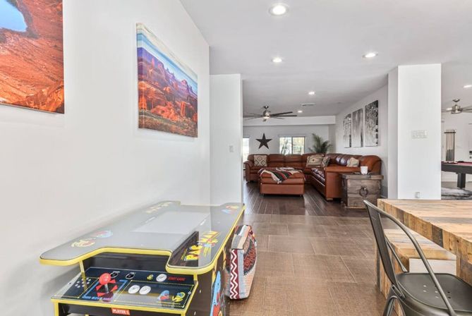 Open-plan living room interior with brown leather sectional, retro arcade game cabinet, rustic wood dining table and metal chairs, desert landscape wall art and star accent — bright modern family entertainment space.