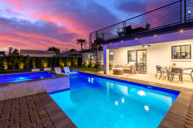 Luxury backyard swimming pool and spa at sunset with glowing blue water, lounge chairs, covered patio seating, outdoor dining table, spiral staircase up to a rooftop deck, palm tree and pink-purple sky.