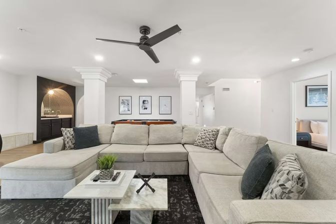 Bright modern home basement entertainment room with oversized light-gray sectional sofa, marble coffee table, black ceiling fan, pool table and built-in wet bar with arched tile backsplash.