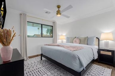Cozy modern bedroom interior with a large bed in light blue linens, pink throw and round accent pillow, two bedside lamps, ceiling fan, patterned rug, potted dried grasses on a dresser, and a window framing a palm tree outside.