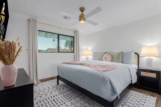 Cozy modern bedroom interior with a large bed in light blue linens, pink throw and round accent pillow, two bedside lamps, ceiling fan, patterned rug, potted dried grasses on a dresser, and a window framing a palm tree outside.
