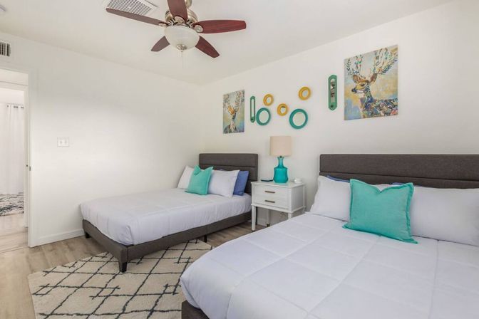 Bright modern guest bedroom with two beds, gray upholstered headboards, white bedding and cheerful teal pillows, white nightstand with turquoise lamp, colorful deer artwork and circular wall decor, ceiling fan and patterned rug.