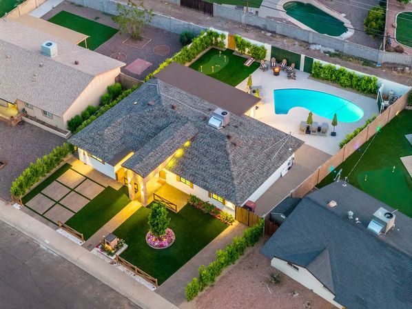 Aerial evening view of a suburban ranch-style home with illuminated front yard and paved lawn squares, backyard kidney-shaped pool, putting green, patio seating and fire pit — backyard oasis in a residential neighborhood.