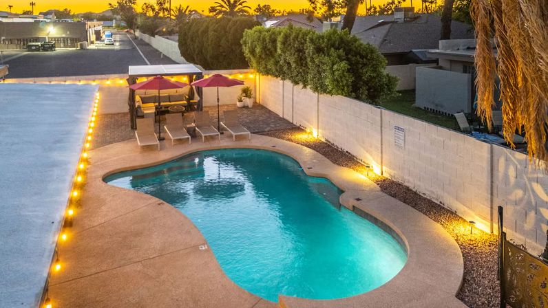 Sunset-lit kidney-shaped backyard pool with turquoise water, illuminated pathway lights, lounge chairs and red umbrellas by a cabana, palm trees and neighborhood rooftops in the background