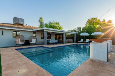 Sunlit suburban backyard with a rectangular swimming pool, covered patio dining area, two wicker egg chairs, lounge chairs and umbrellas, and decorative water bowls at sunset.