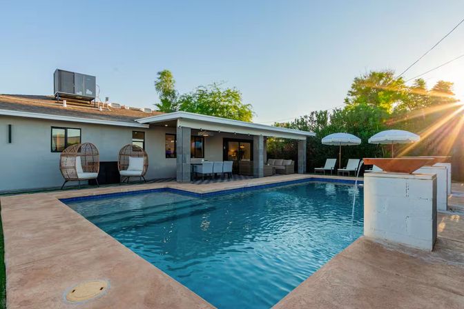 Sunlit suburban backyard with a rectangular swimming pool, covered patio dining area, two wicker egg chairs, lounge chairs and umbrellas, and decorative water bowls at sunset.