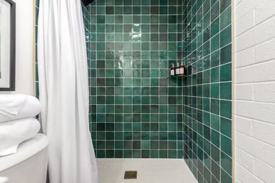 Walk-in bathroom shower with glossy dark green square tiles, white textured shower curtain, white mosaic floor with brass drain, stacked towels on tub edge and corner shelf holding pump bottles
