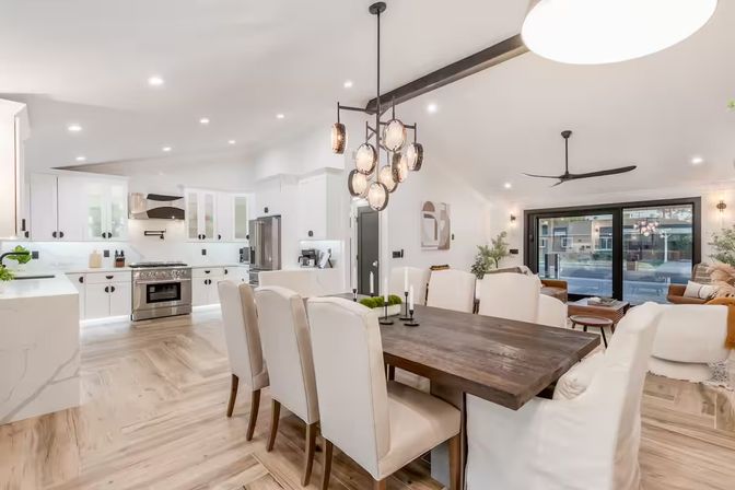 Bright open-concept kitchen and dining room with white cabinetry and marble island, stainless steel range, dark wood dining table with cream upholstered chairs, modern pendant lights, light herringbone wood floors, and large sliding glass doors to a patio.
