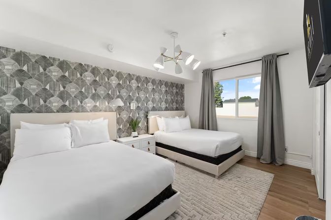 Bright modern hotel bedroom with two queen beds and white linens, geometric gray accent wall, bedside table with lamp and plant, window with gray curtains and hardwood floors.
