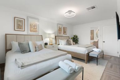 Bright modern guest bedroom with two queen beds, neutral linens, bedside lamp, rolled spa towels on benches, framed botanical art and a potted plant.